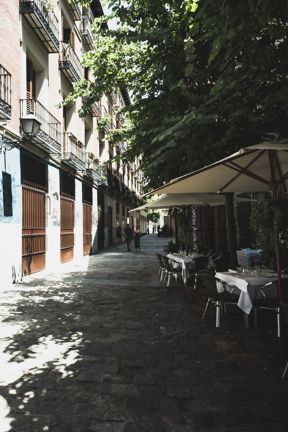 Shaded cobblestone alley with outdoor restaurant seating under umbrella and lush trees in Madrid.