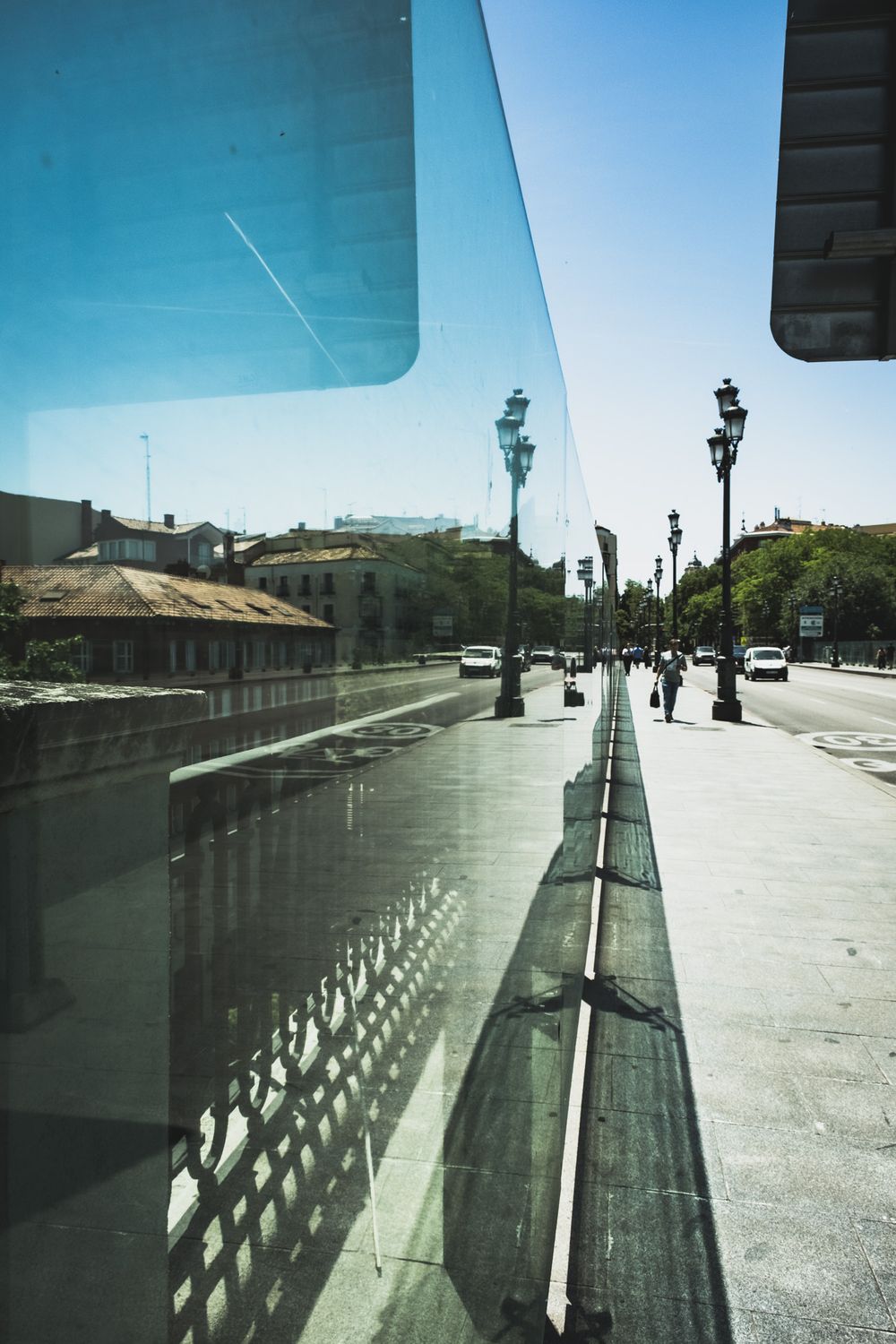 City street reflected in modern glass building facade with ornate lamp posts under clear blue sky.