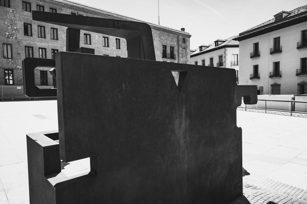 Black and white photo of abstract metal sculpture in open plaza with historic buildings behind.