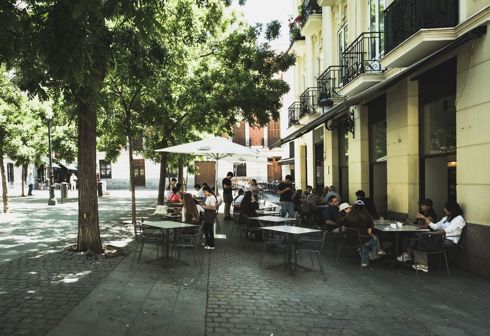 Outdoor cafe terrace with people dining under trees on a cobblestone street in Madrid, Spain.