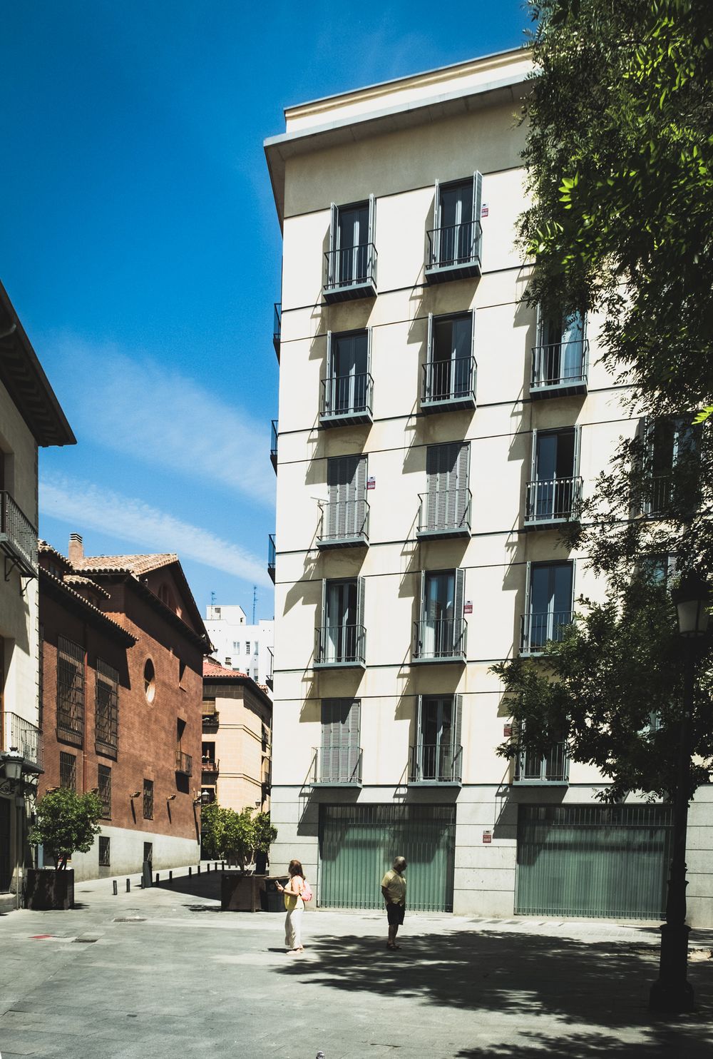 Tall white residential building with balconies on sunny Madrid street with pedestrians walking below.