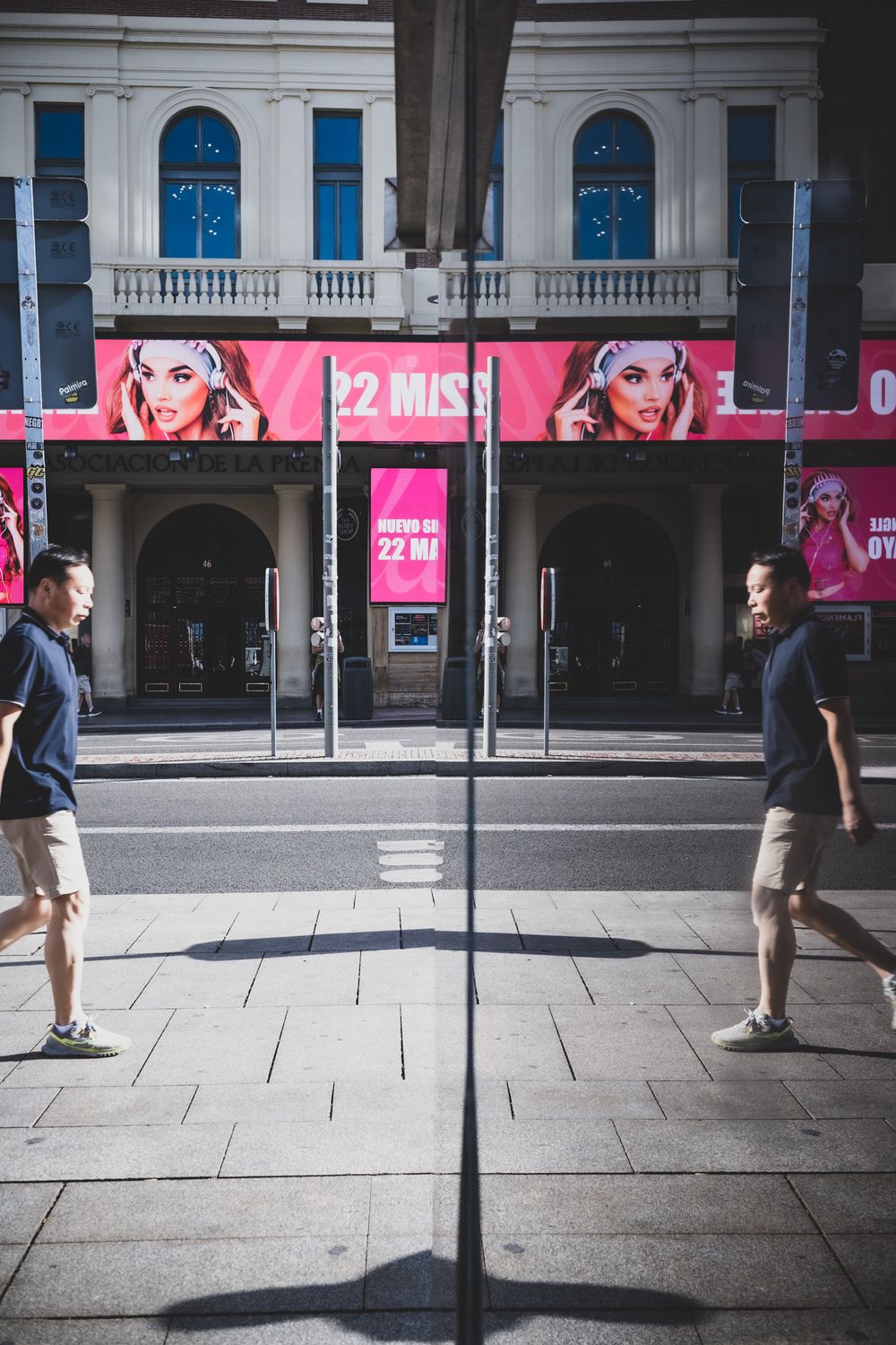 Man reflected in glass panel on urban sidewalk with pink theater marquee in background.