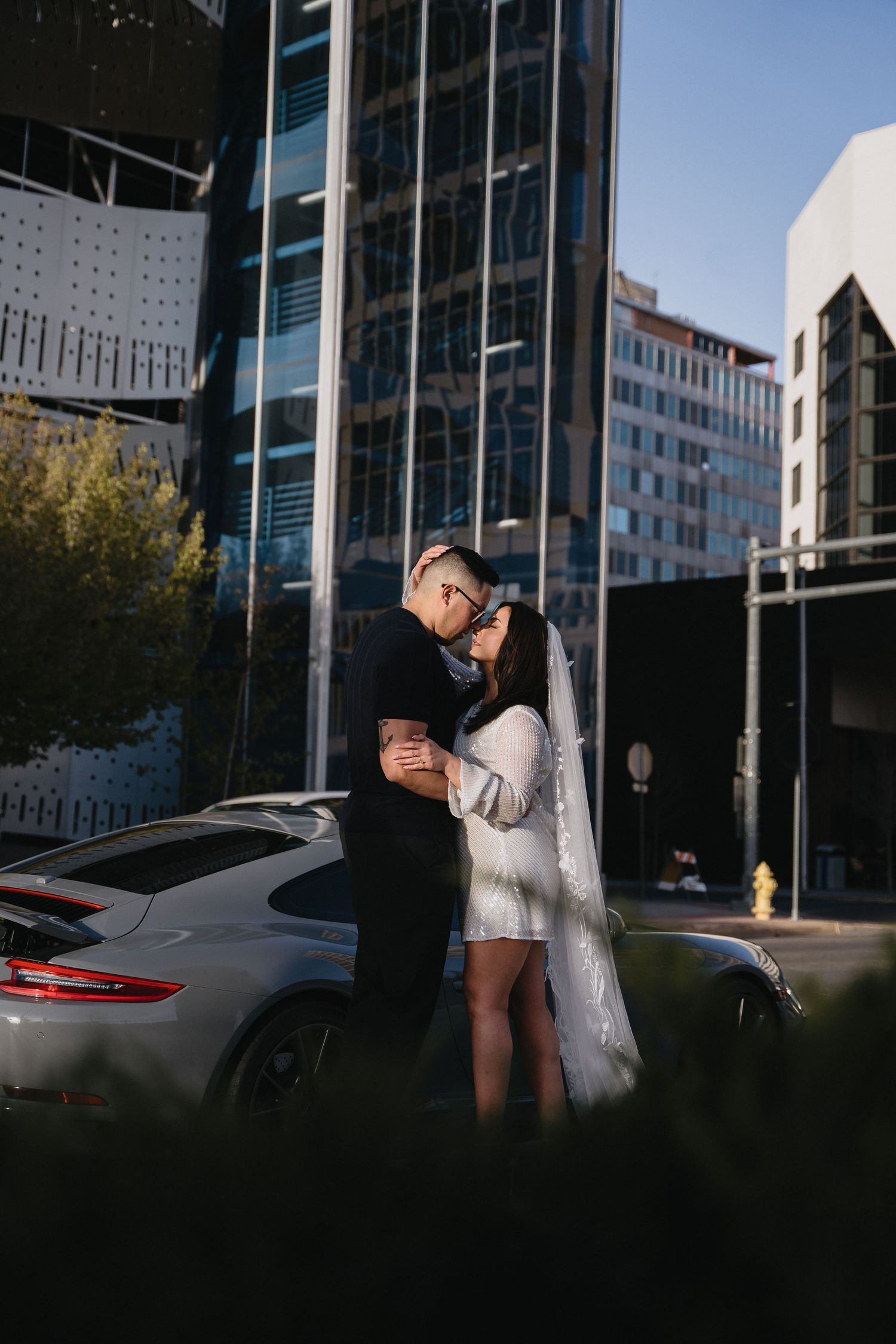 Bride in short white dress and veil kissing groom in navy suit beside a Porsche in a city setting in Albuquerque New Mexico