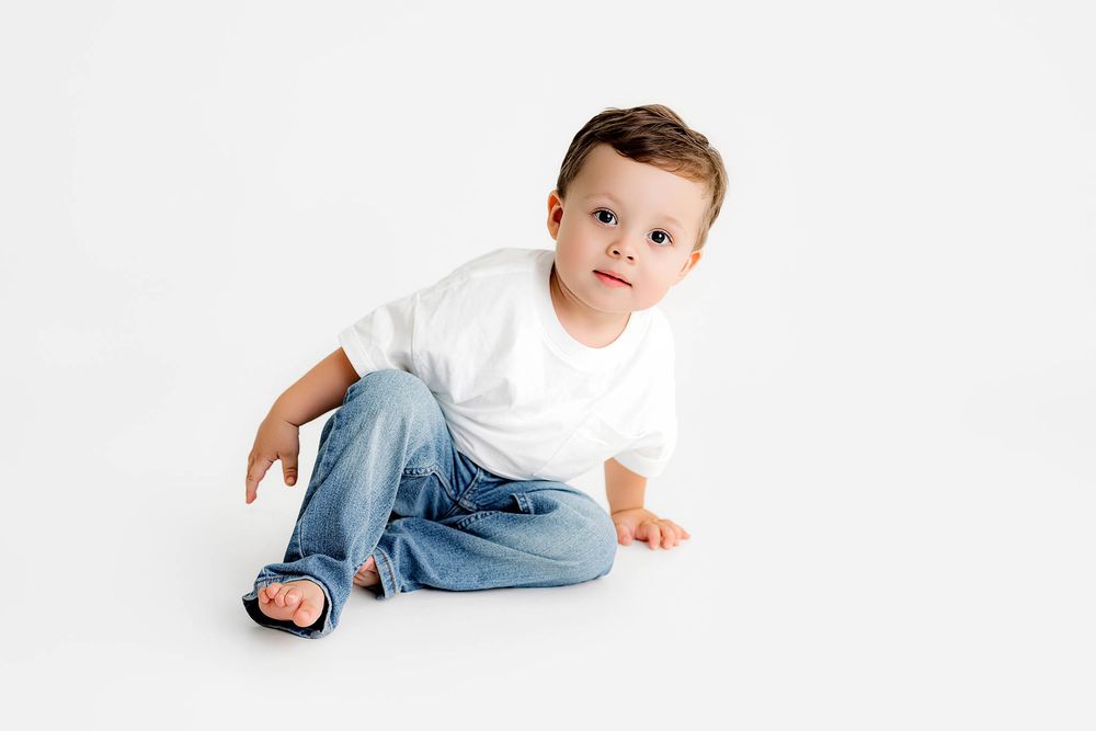 Cute toddler boy in white t-shirt and jeans sitting on white background, looking at camera.