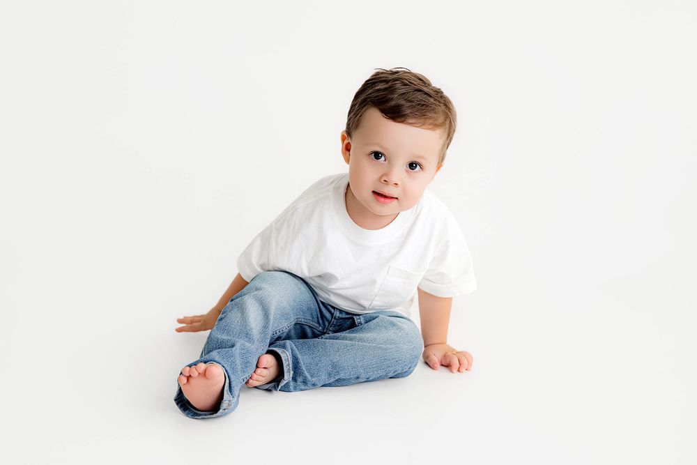 Cute toddler boy in white t-shirt and jeans sitting on a white studio floor.