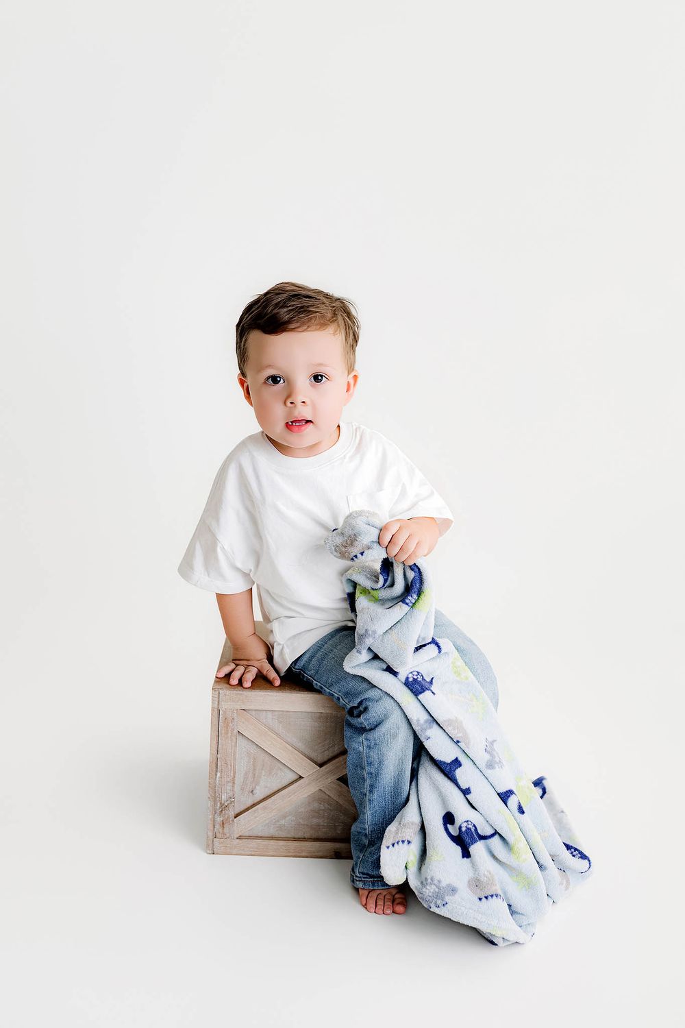 Toddler boy in white t-shirt and jeans sits on wooden crate holding a dinosaur print blanket on white background.