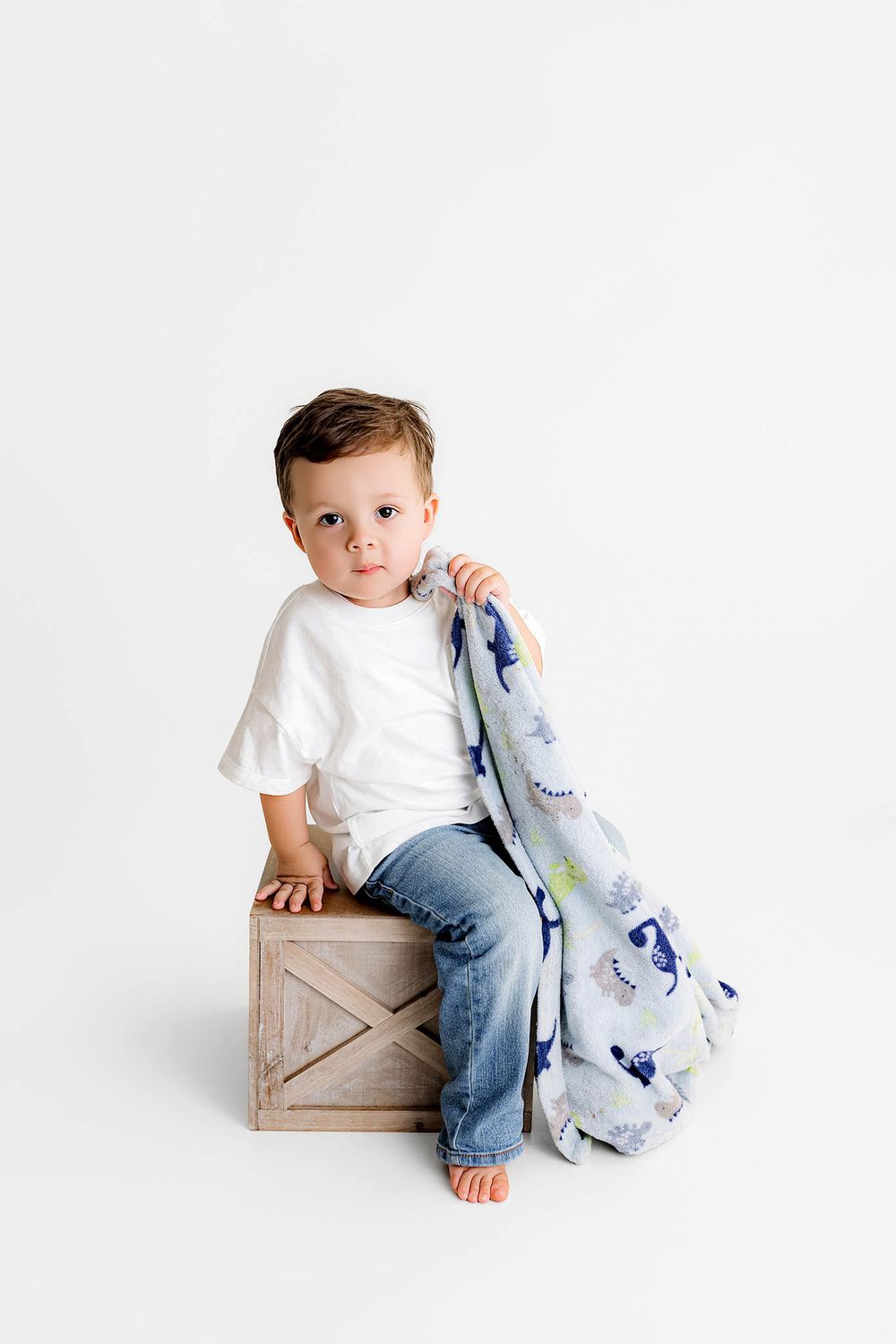 Toddler boy in white tee and jeans sits on wooden crate holding a blue dinosaur print blanket against white background.
