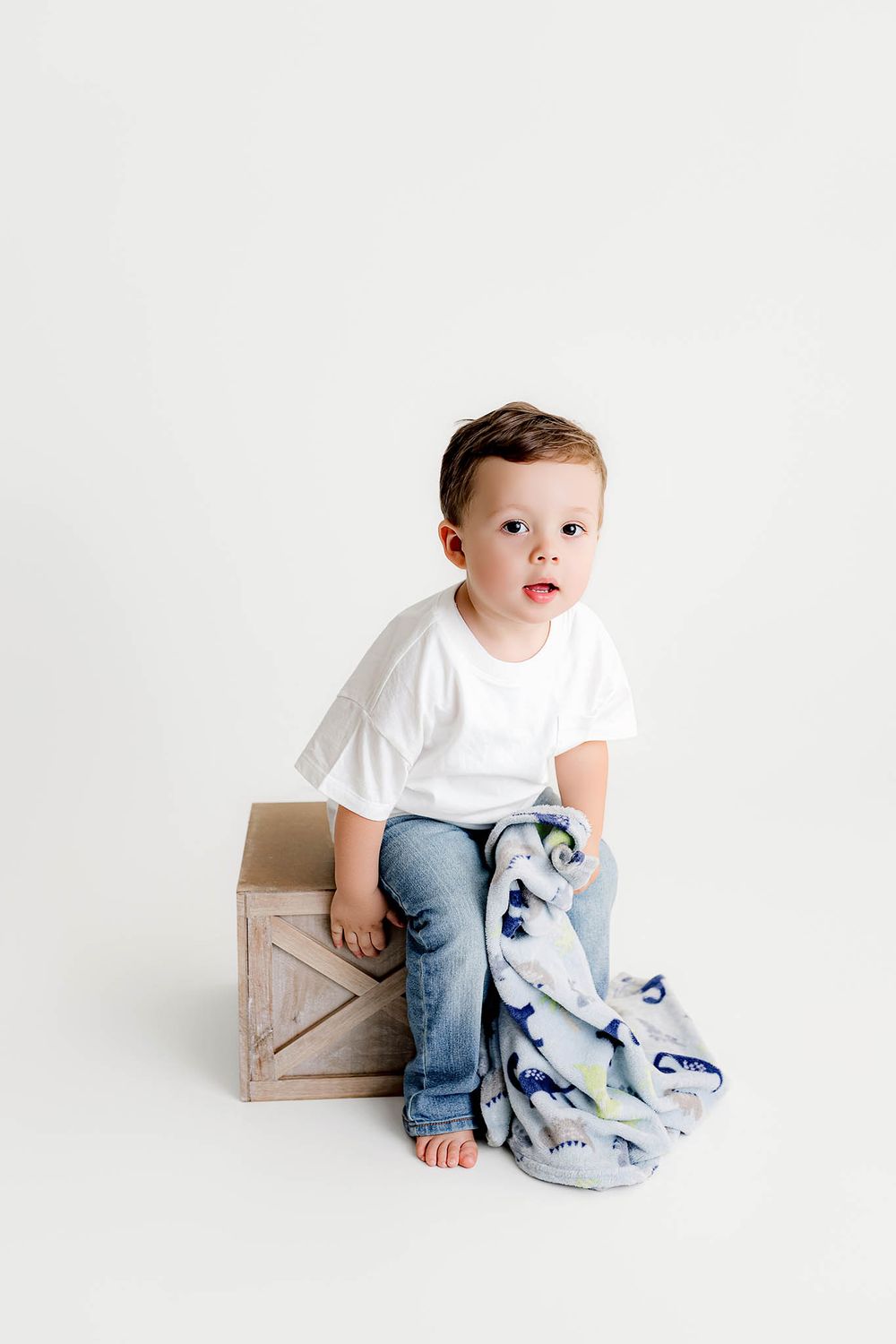 Young boy in white t-shirt and jeans sits on rustic wooden crate with colorful dinosaur blanket in white studio.