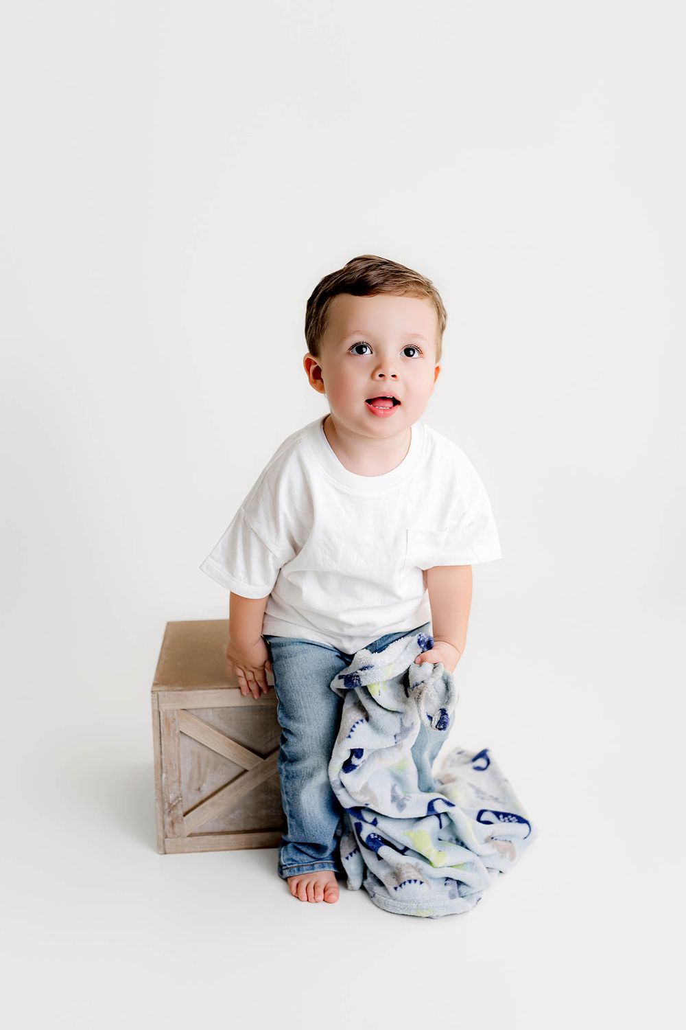 Cute toddler boy in white tee and jeans holding a blanket while standing next to a wooden crate.