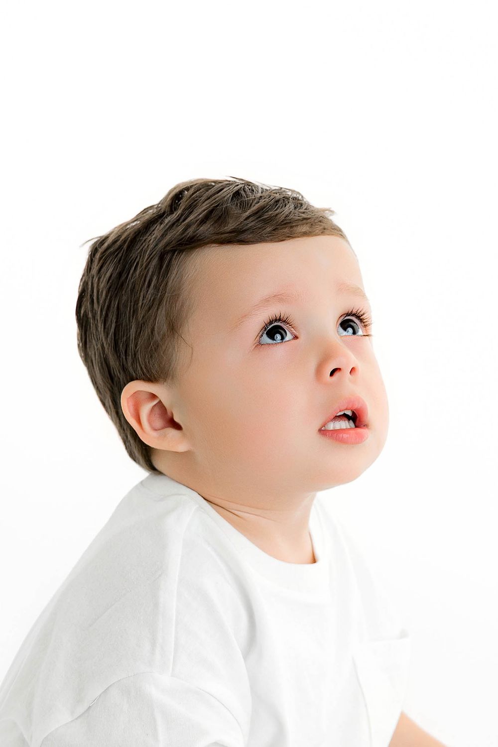 Curious toddler boy with blue eyes looking up, wearing a white t-shirt against a white background.