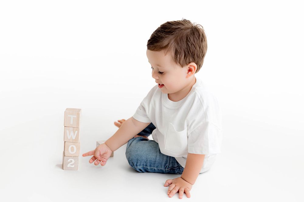 Toddler boy in white shirt and jeans playing with stacked wooden number blocks on white background.