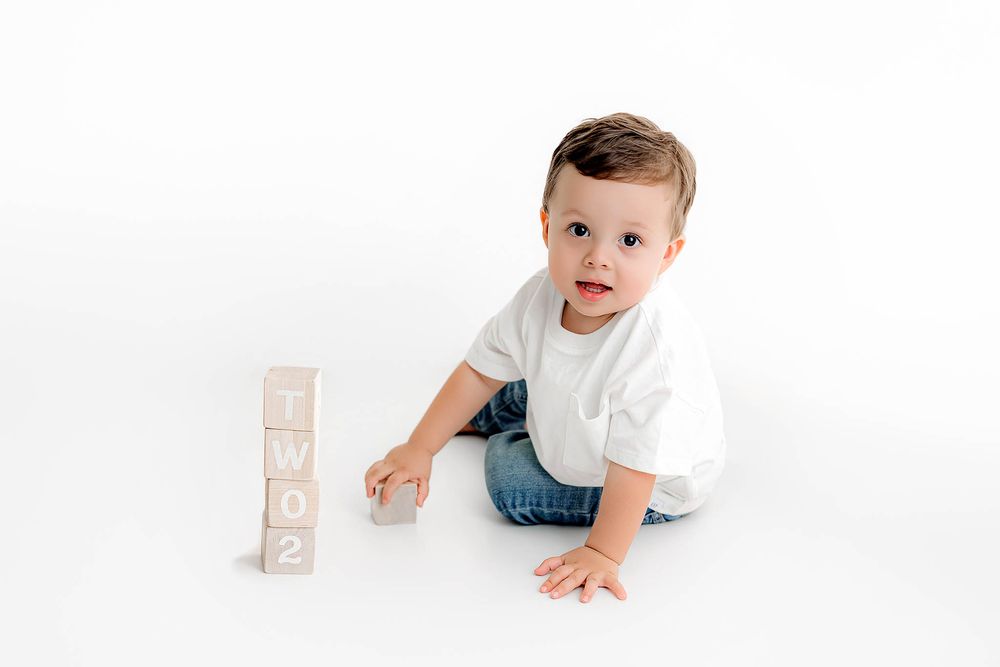 Adorable toddler boy in white tee and jeans sitting beside stacked wooden number blocks smiling.