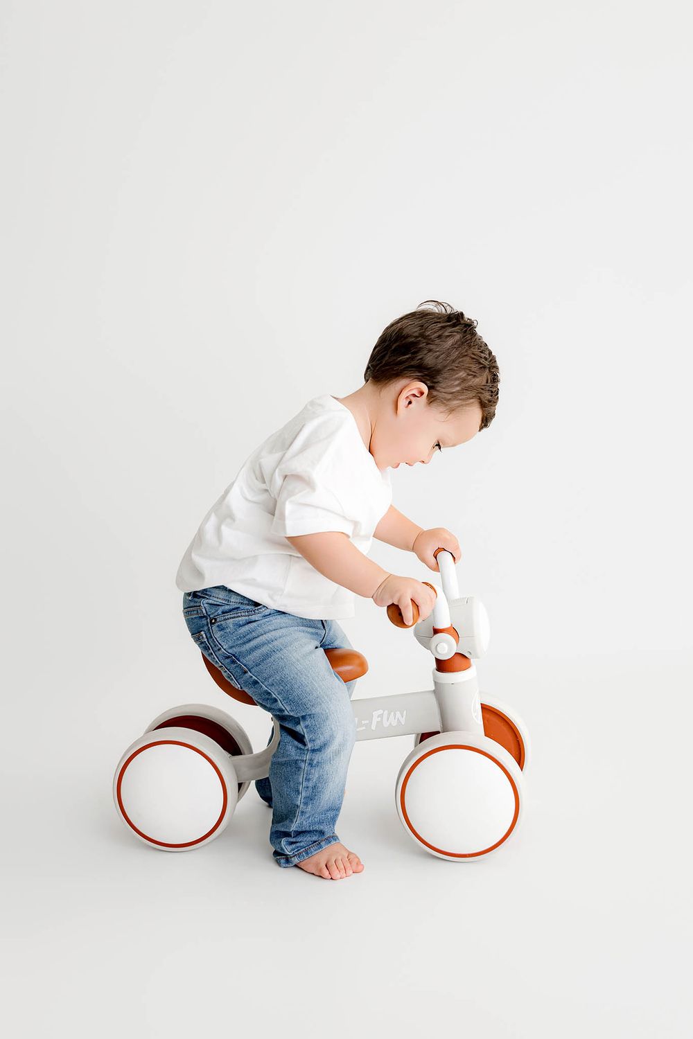 Two year old boy's second birthday photos in white shirt and jeans riding a small balance bike on a white background.