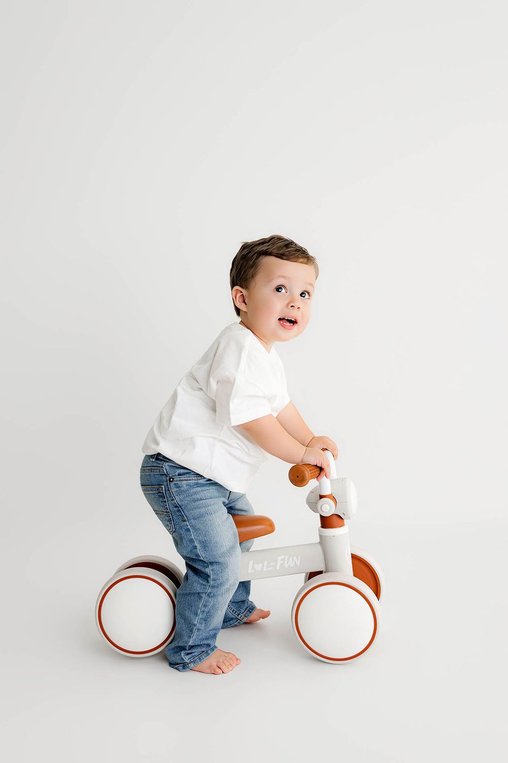 Toddler boy in white tee and jeans riding a white and brown balance bike on a white background.