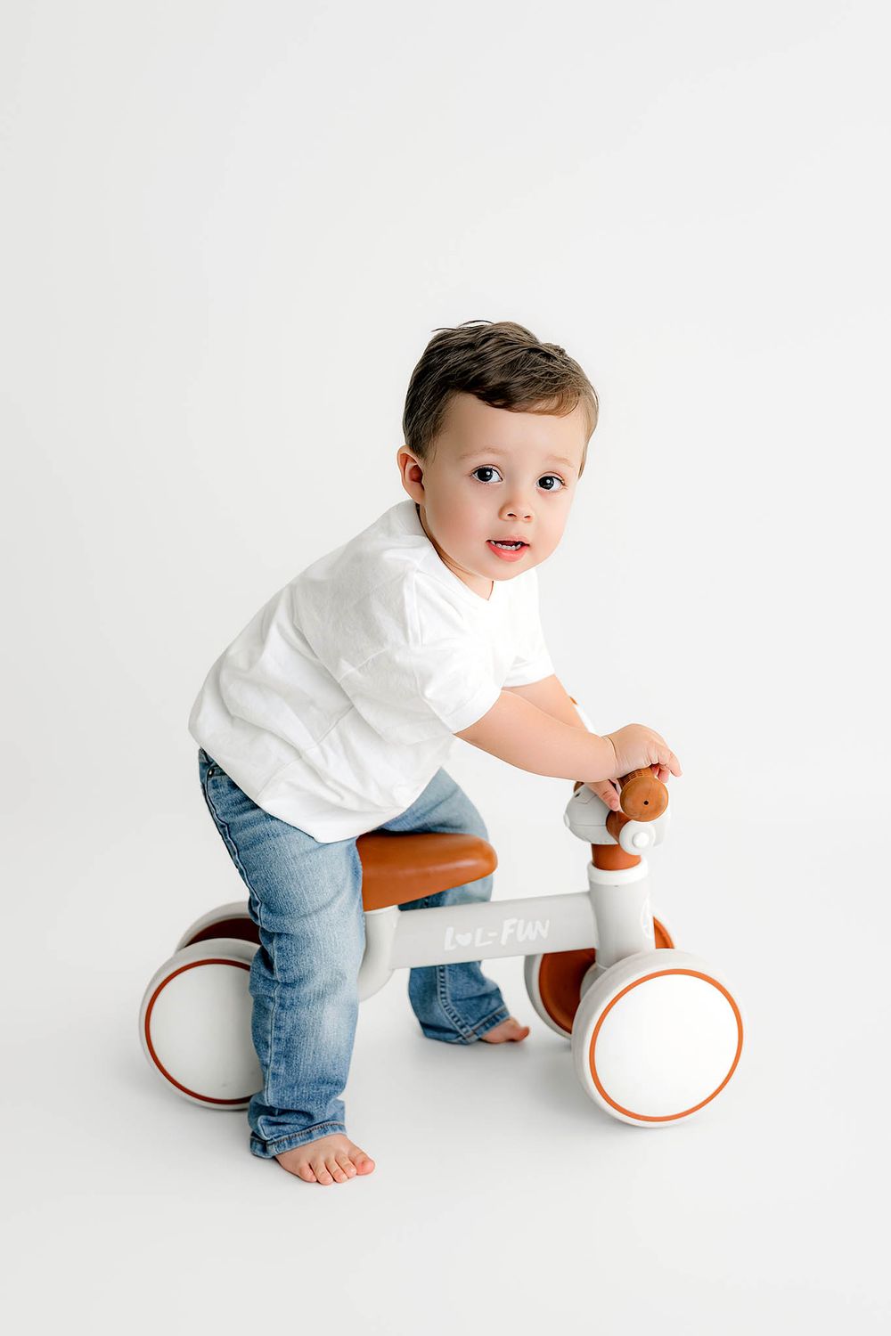 Two year old boy in white shirt and jeans riding a white balance bike toy on in a bright white studio for second birthday photos.