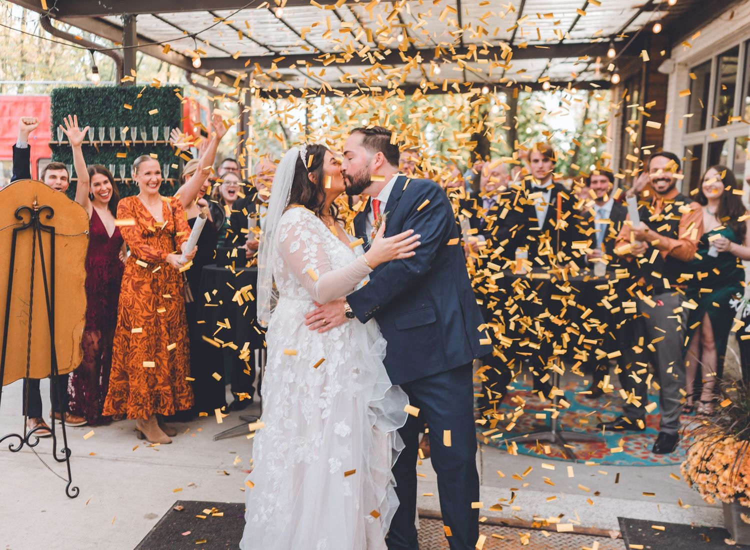 Newlyweds share a kiss surrounded by golden confetti while guests celebrate at an outdoor wedding reception.