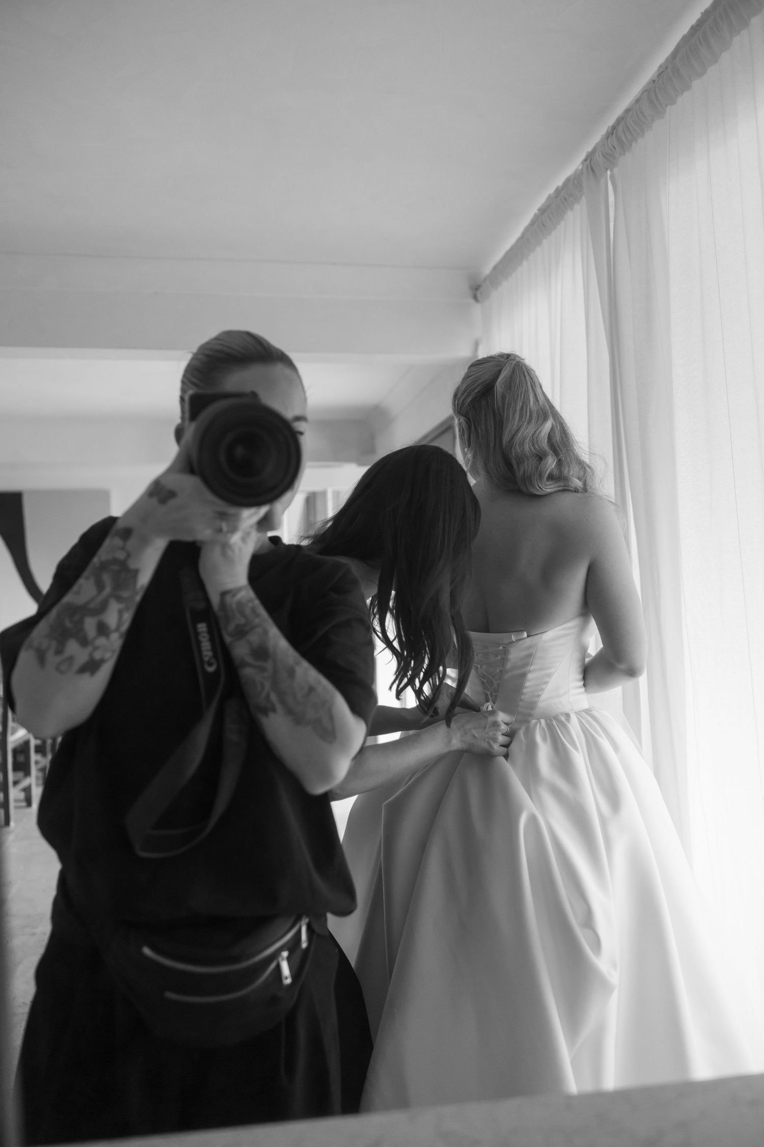 Black and white photo of tattooed photographer capturing bride getting dress fastened by assistant near window.