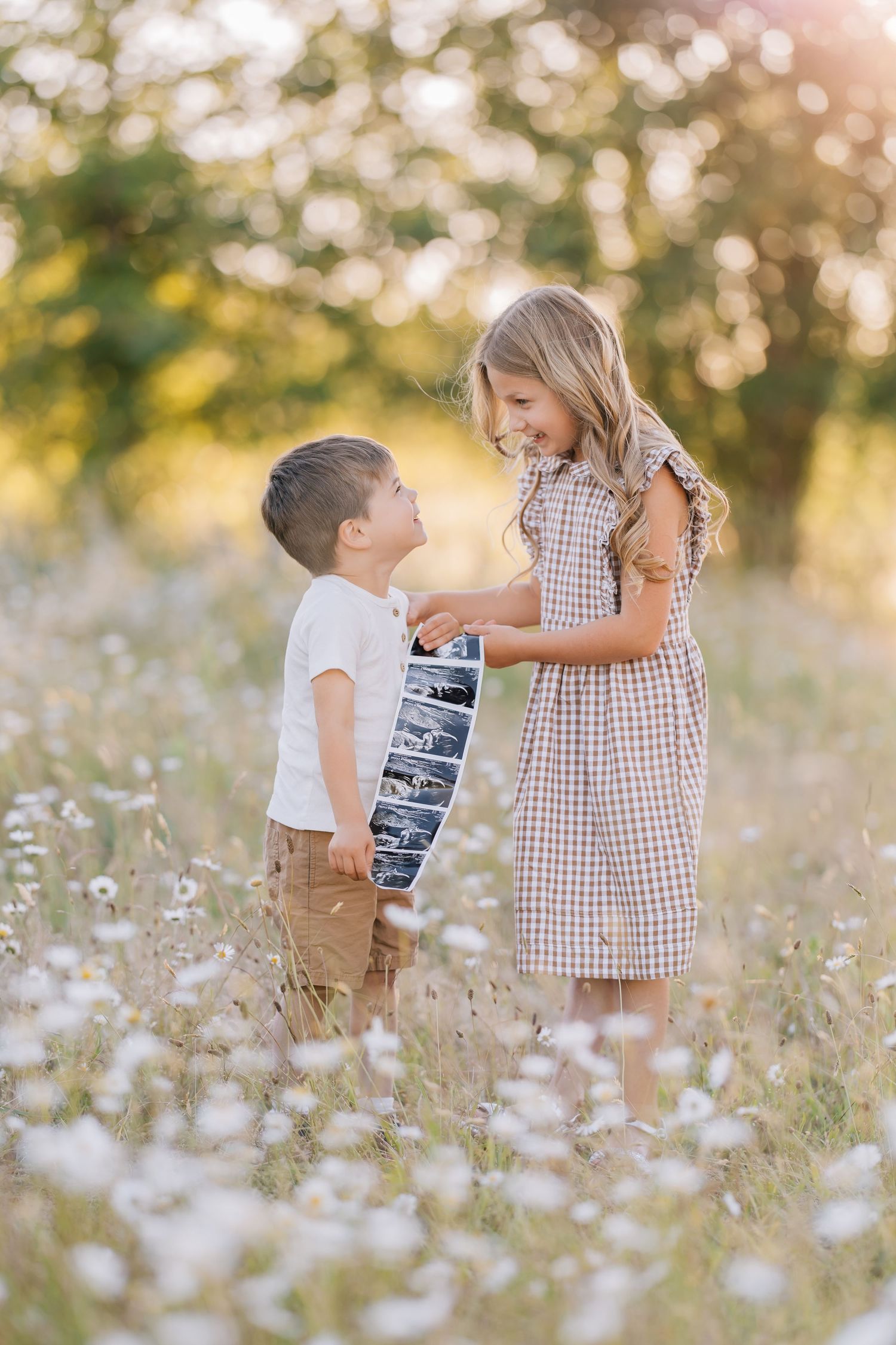 Two children share a joyful moment holding ultrasound photos in a sunny daisy field.