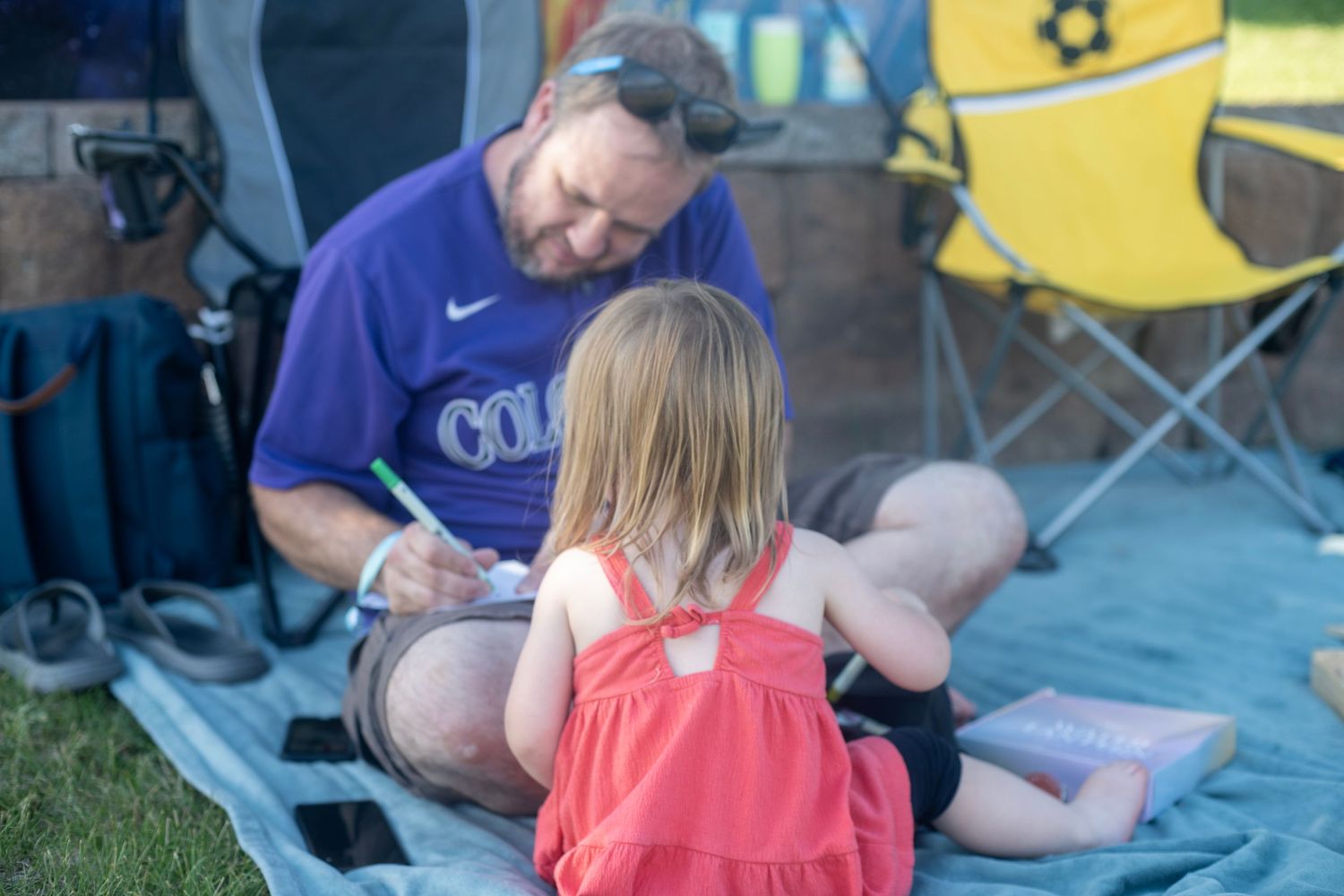 A man in a purple Colorado shirt sits on a blanket outdoors writing with a young girl in a red dress.