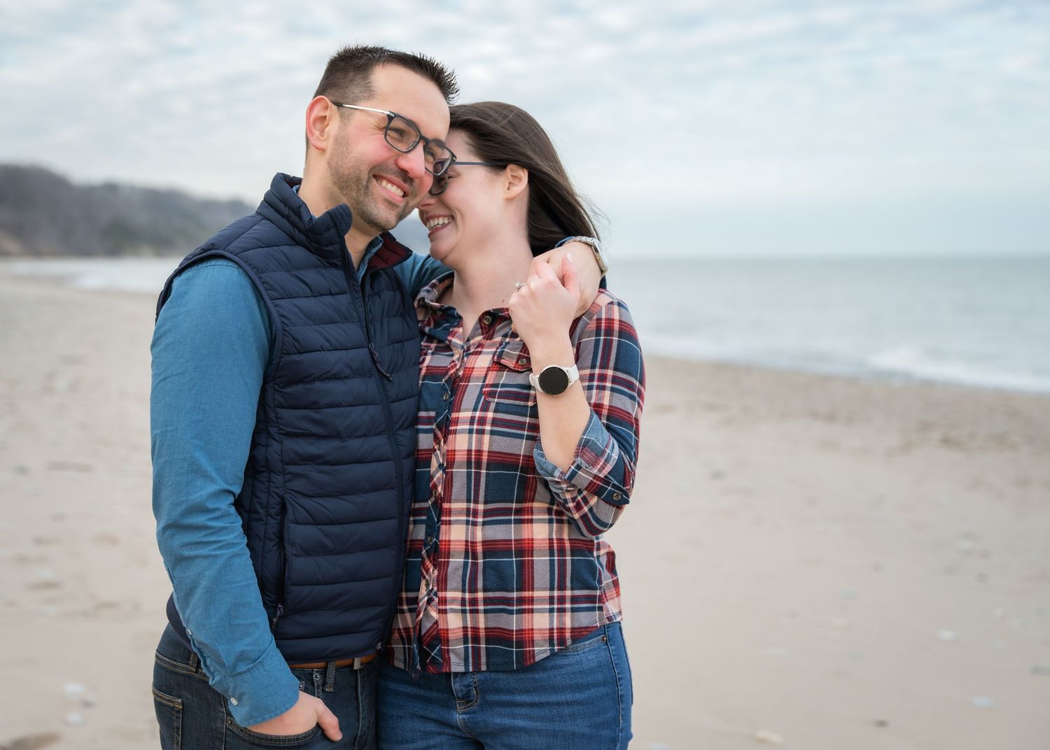 Couple smiling and embracing on a sandy beach, wearing casual flannel and vest, with ocean waves in the background.