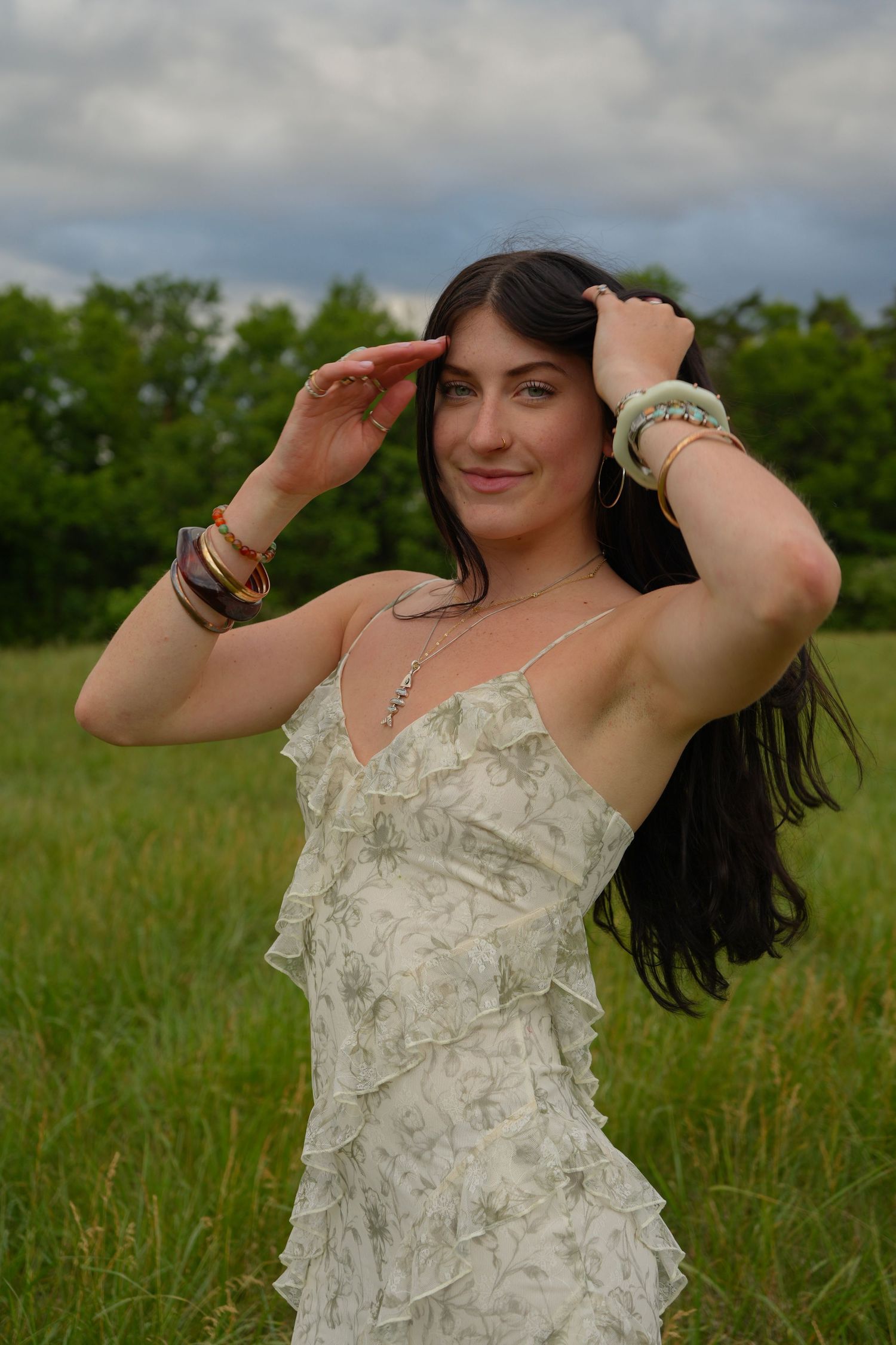 Young brunette woman in white floral sundress posing in green meadow under cloudy sky.