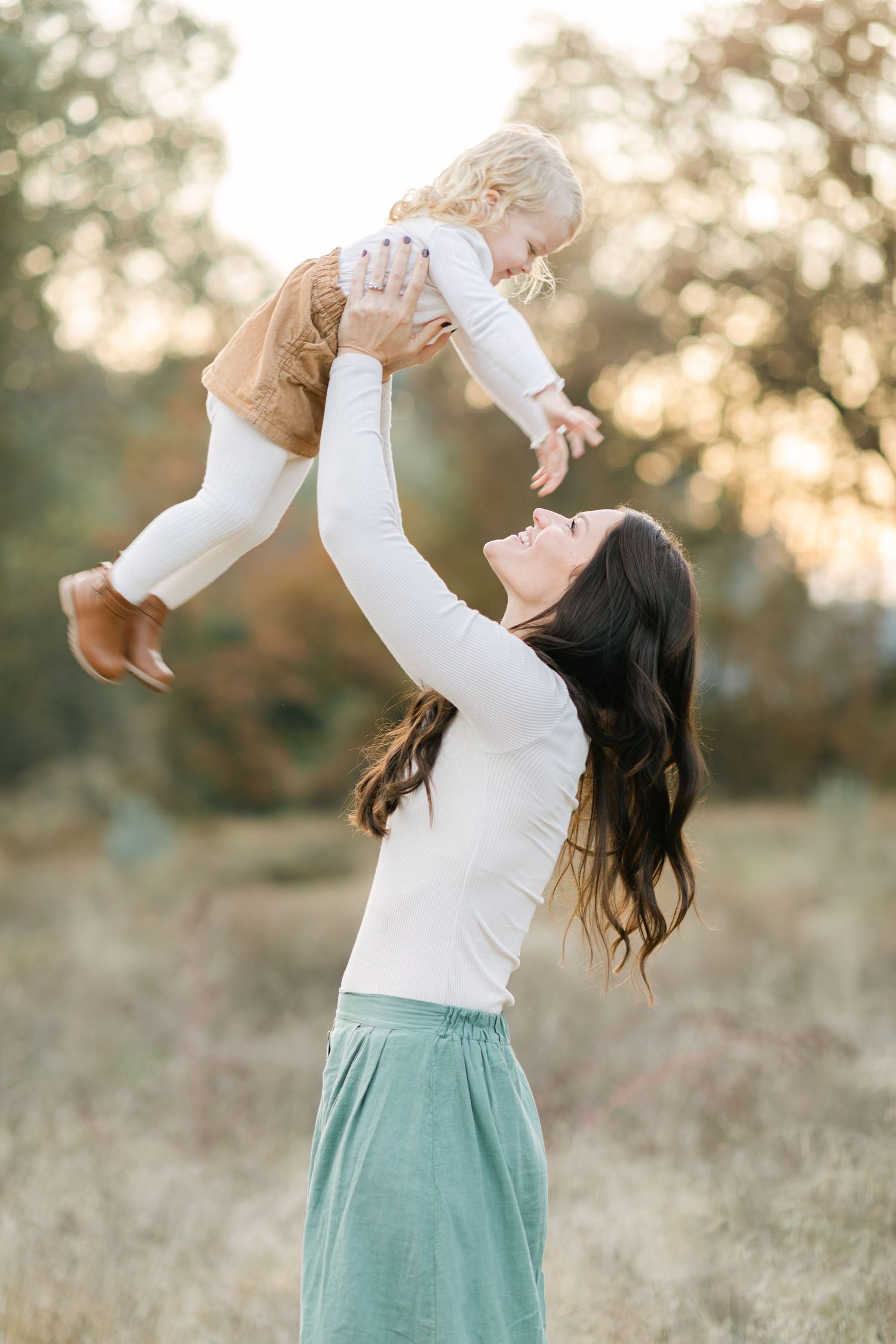 A joyful mother lifts her smiling toddler high in a sunlit autumn field, both wearing white tops.