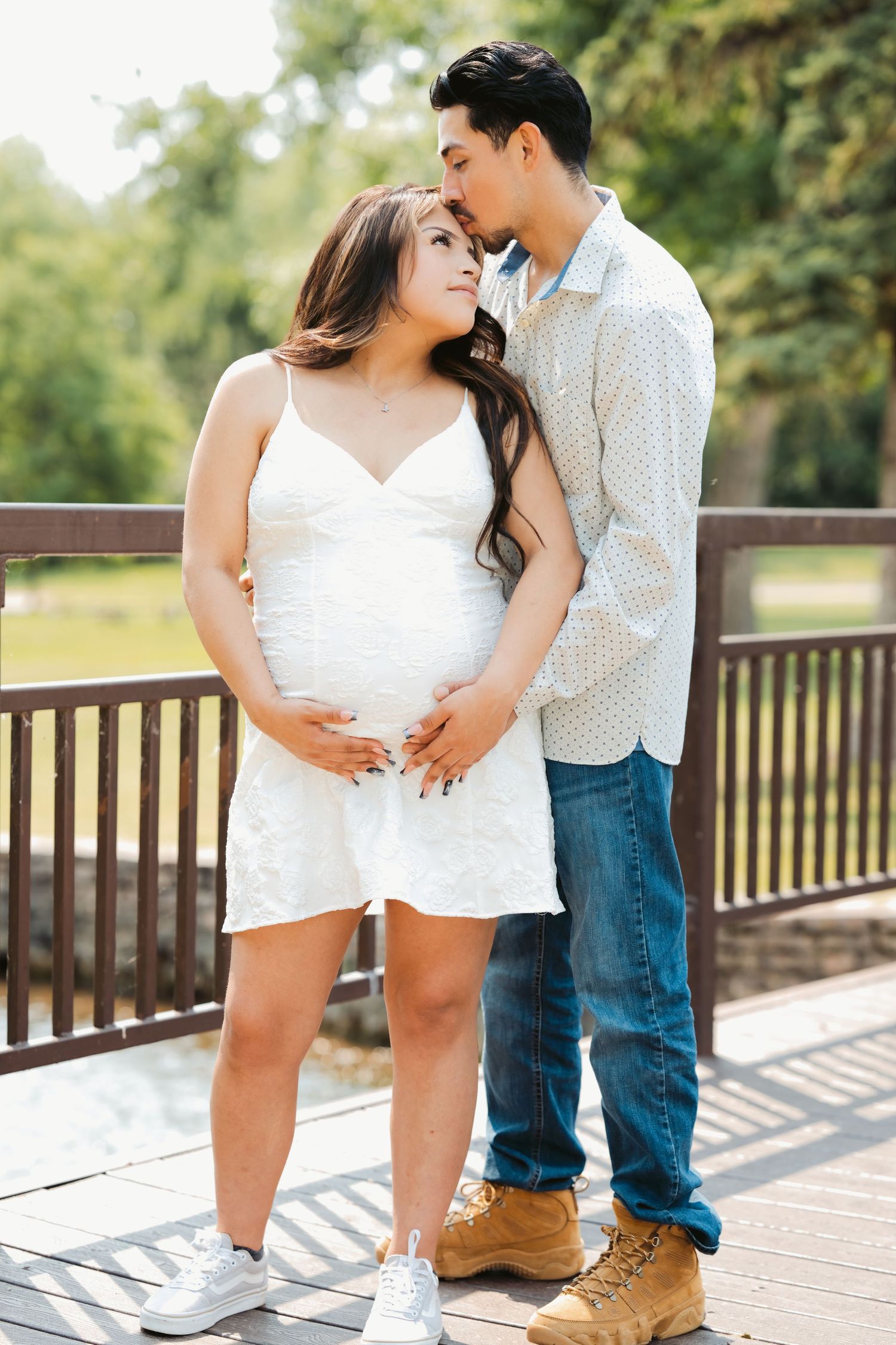 Couple sharing a tender moment on a wooden bridge, pregnant woman in white dress cradling her baby bump outdoors.