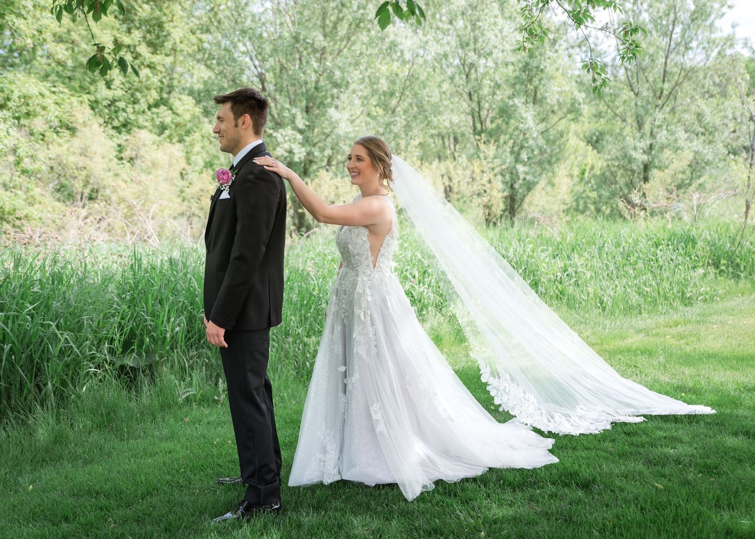 Bride in lace gown adjusts groom's suit outdoors near lush greenery on their wedding day.