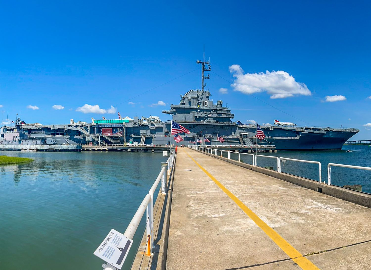A large naval warship docked at a pier under a bright blue sky with fluffy white clouds on a sunny day.