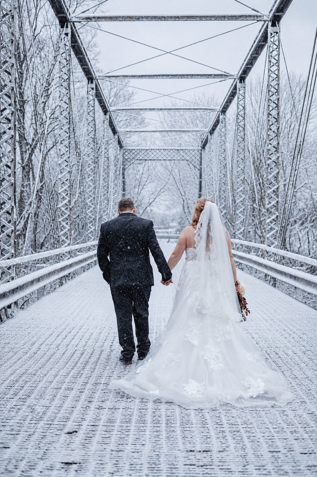 Bride and groom walk hand-in-hand across a snow-covered iron bridge during a winter wedding snowstorm.