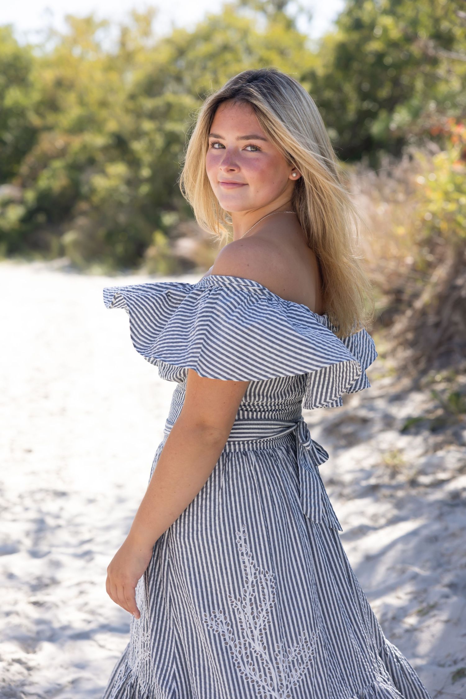 Beautiful blonde woman in striped off-shoulder dress posing on sandy beach path in sunlight.