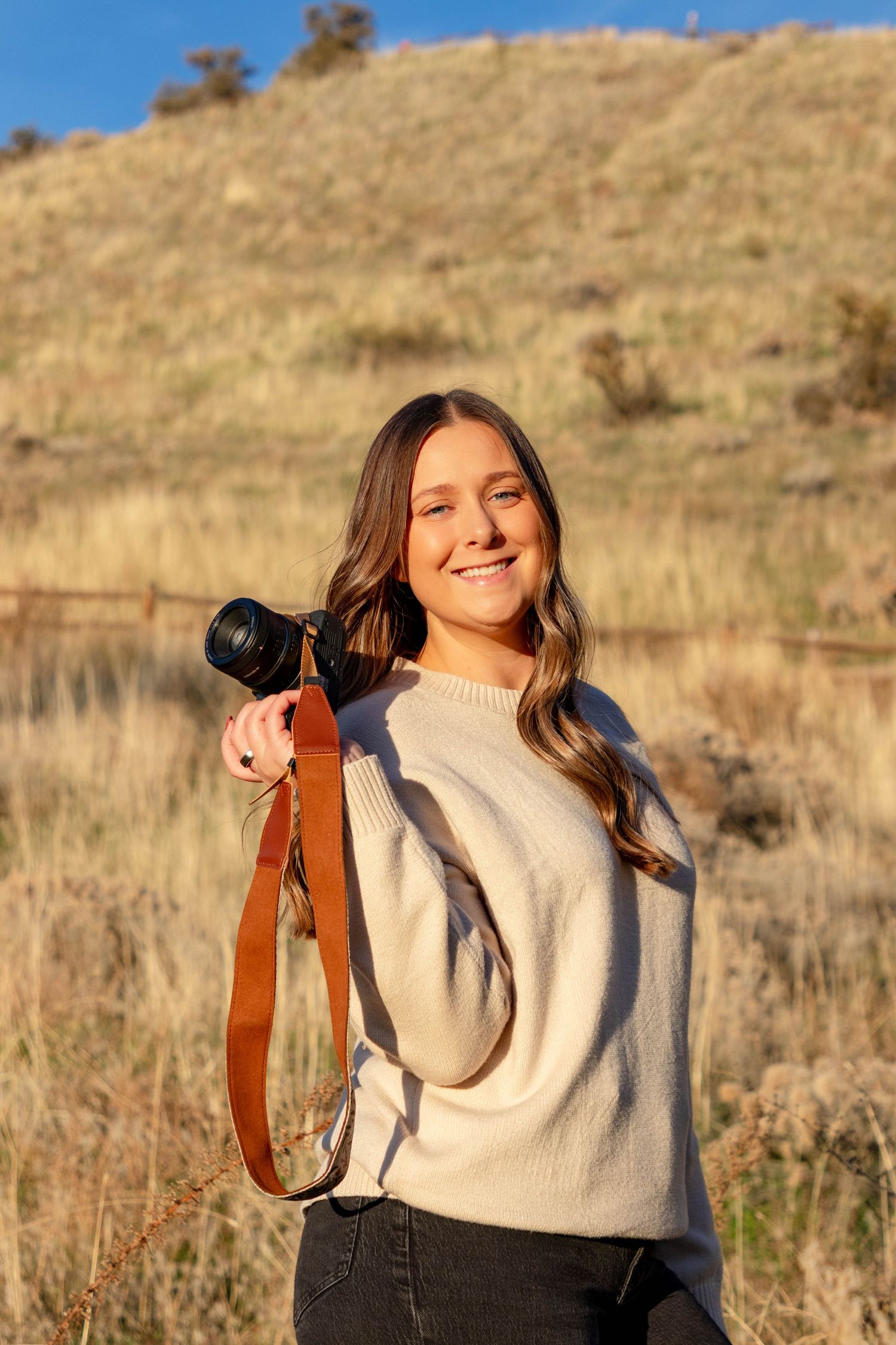 Smiling young woman holding a DSLR camera with brown strap, wearing cream sweater outdoors in golden field.