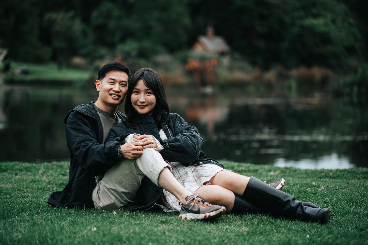 A young Asian couple sitting together on green grass by a serene lake, smiling warmly taken by Melbourne engagement photographer.