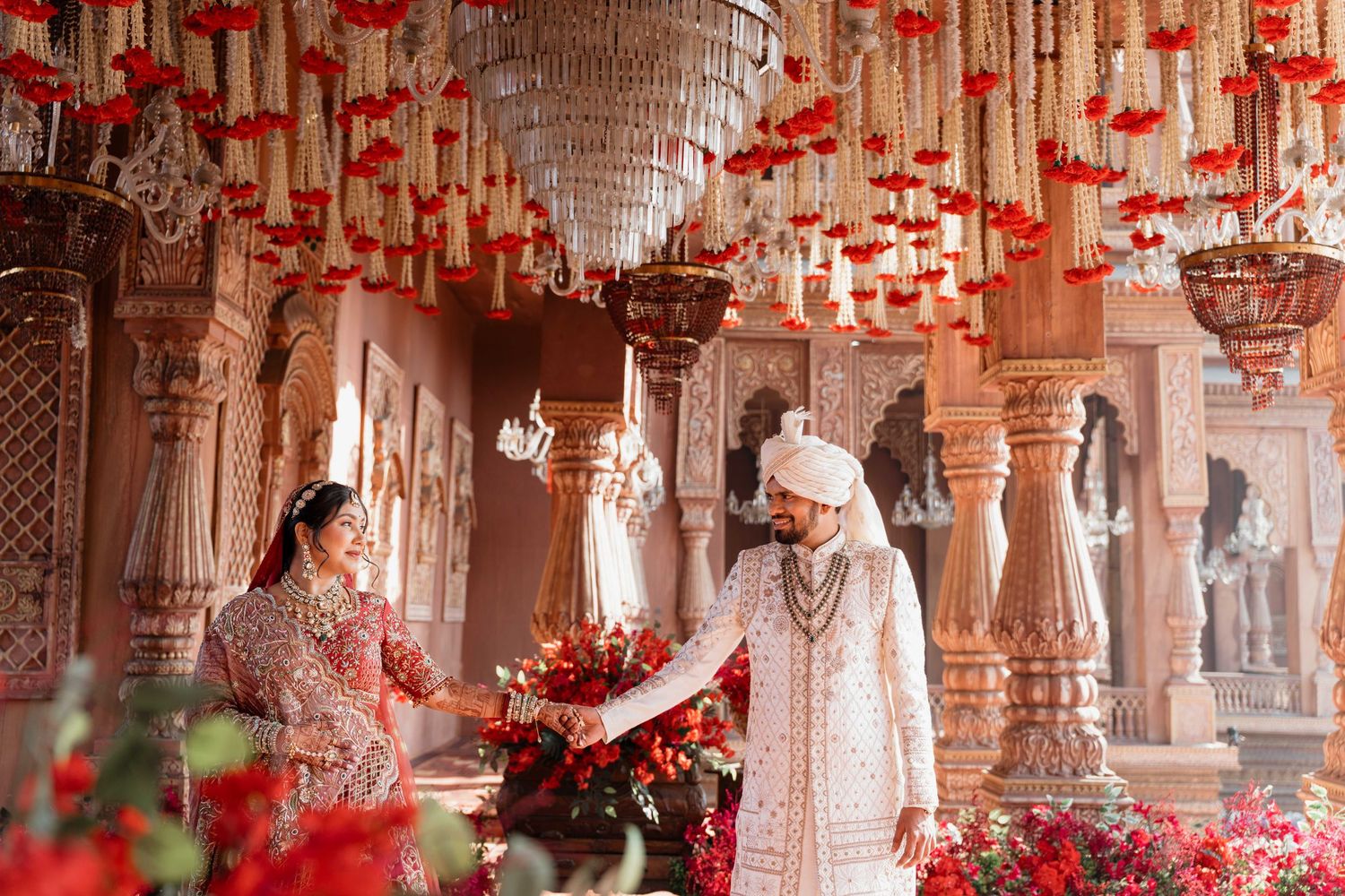Indian couple in traditional wedding attire standing in ornate temple adorned with red floral decorations and chandelier.