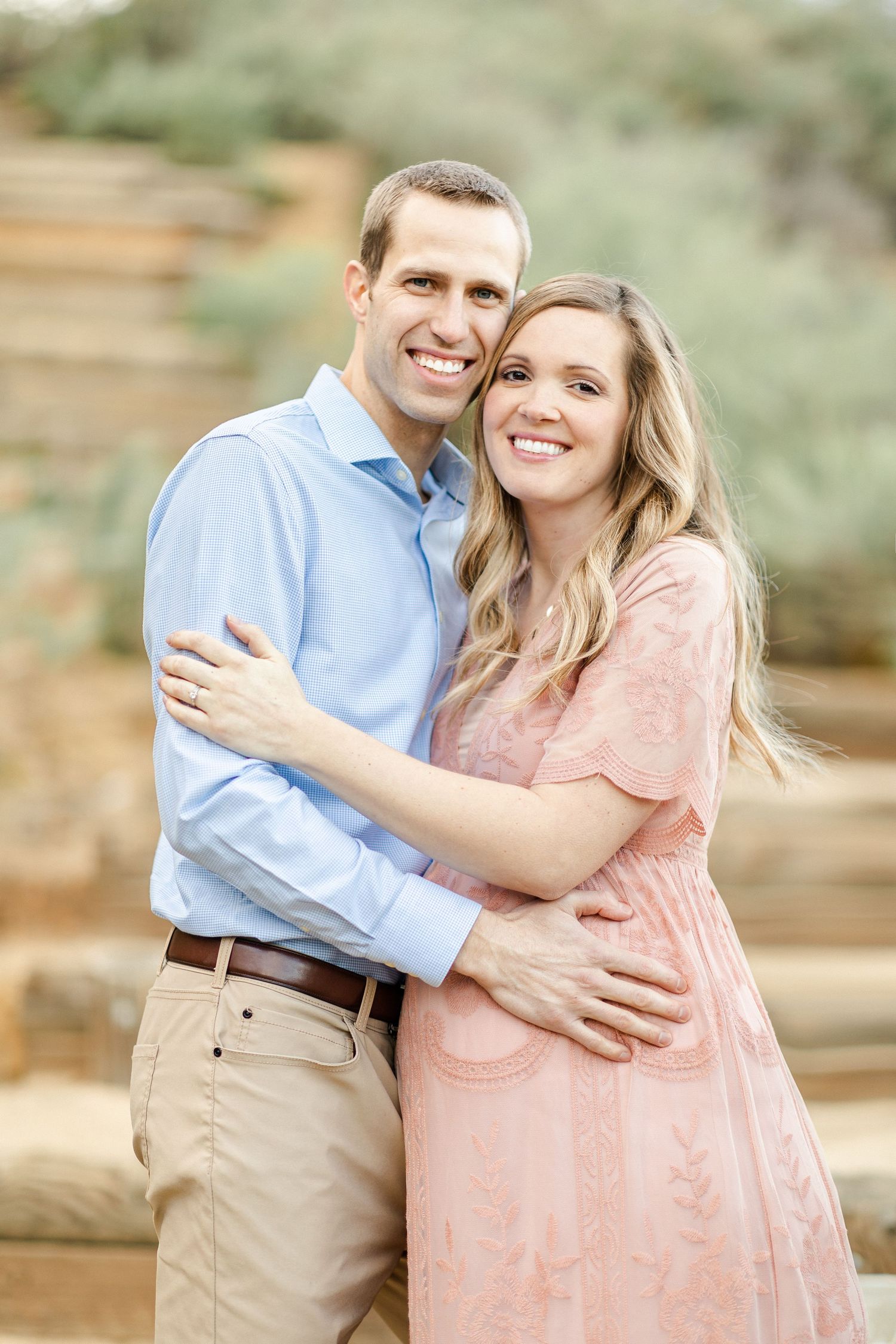 A smiling couple embraces outdoors, man in blue shirt and khakis, woman in pink dress, with stone steps behind them.