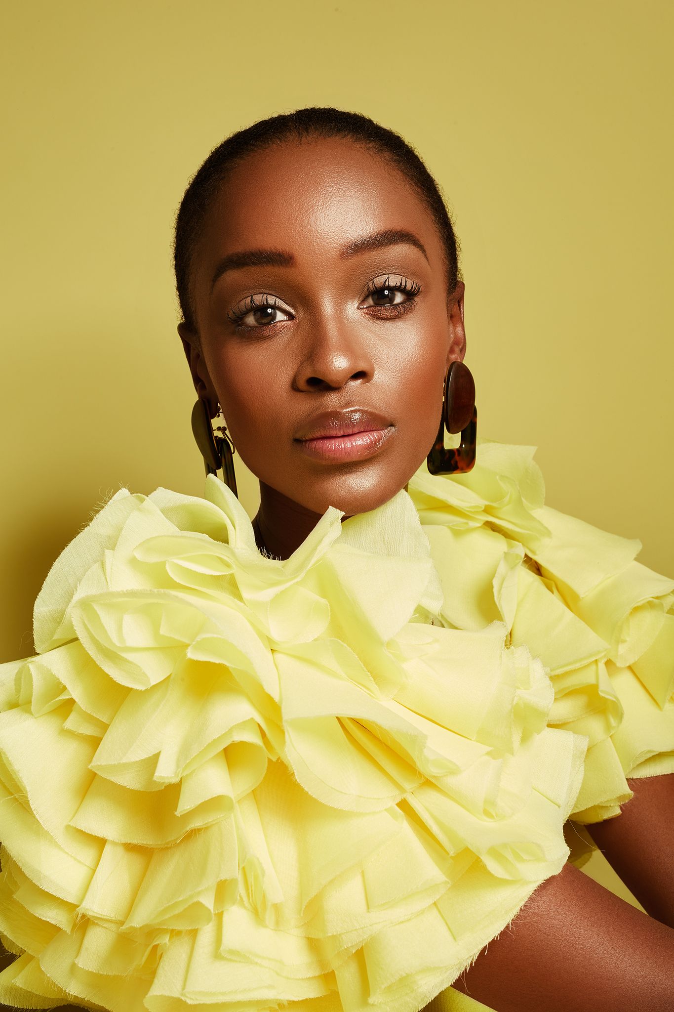 Close-up portrait of a Black woman in a yellow ruffled dress with gold earrings on a yellow background.