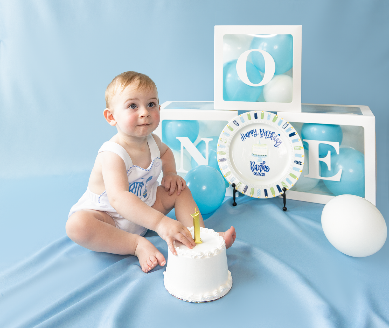 Baby boy celebrating first birthday sitting with white cake in front of letter blocks on blue backdrop.