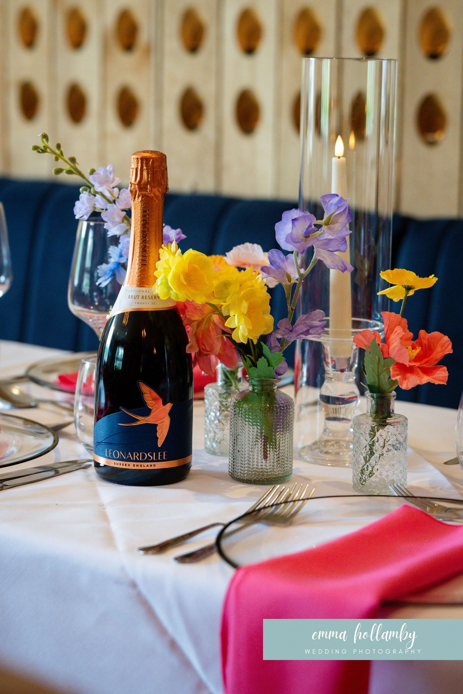 Elegant wedding table setting with colorful flowers, champagne bottle, candle, and pink napkins.