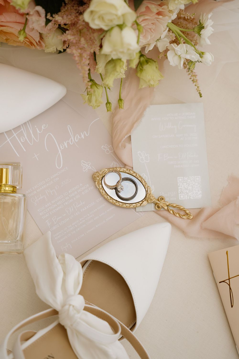 Elegant wedding flat lay with blush invitation, gold mirror, rings, white heels, perfume, and pink floral bouquet.