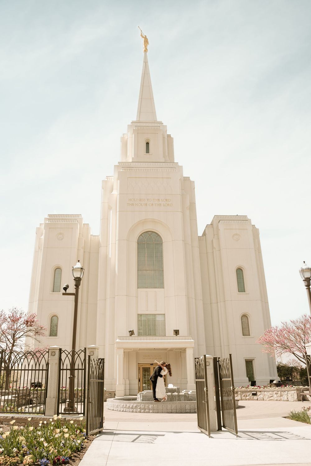 Bride and groom kissing on steps of white LDS temple with gold spire, blooming trees, and iron gates in spring.