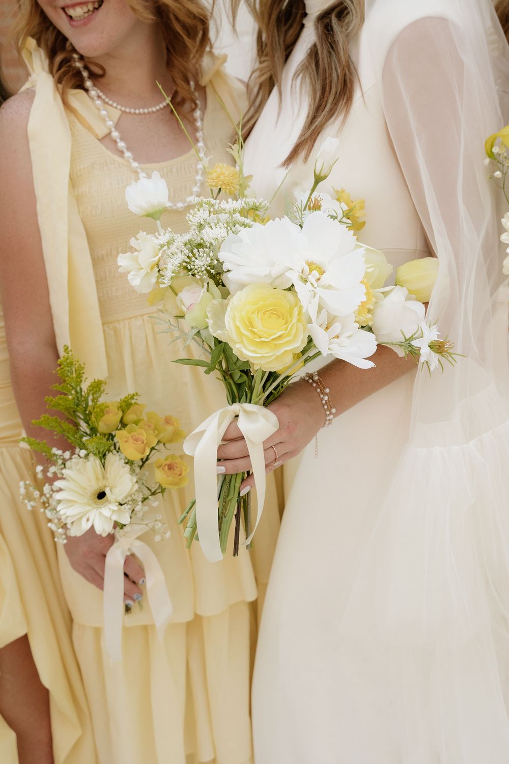 Bride and bridesmaid holding yellow and white floral bouquets tied with ribbon, wearing yellow and white wedding attire.