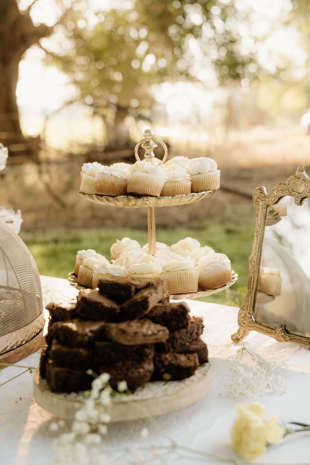 Elegant tiered dessert stand with white frosted cupcakes and chocolate brownies at an outdoor wedding reception.
