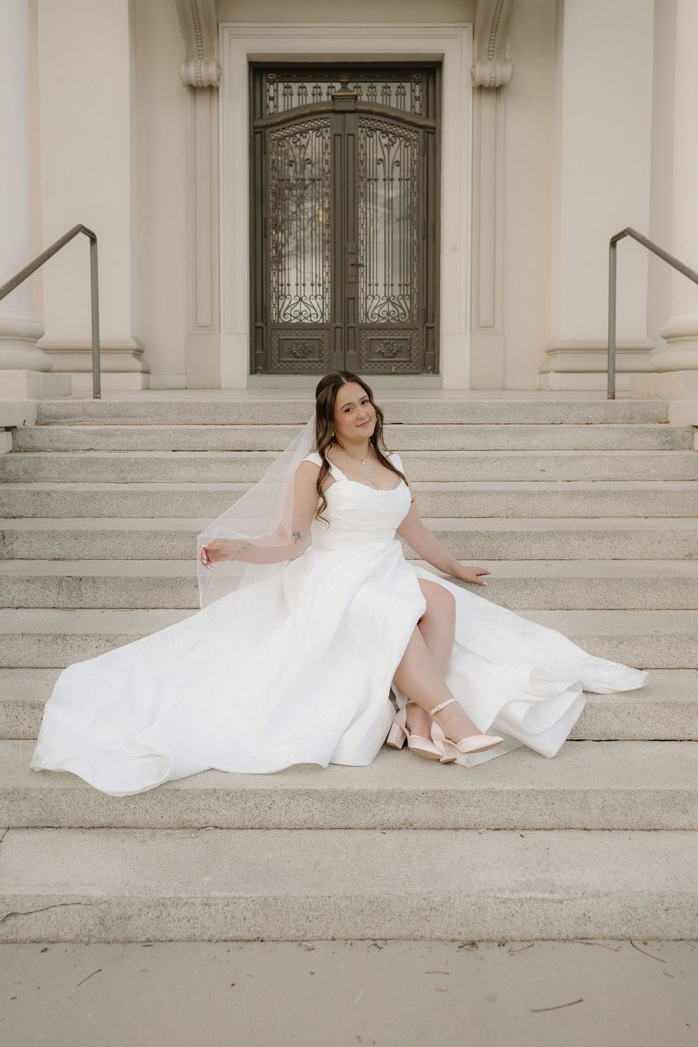 Bride in white gown with veil sits gracefully on stone steps before ornate doors.