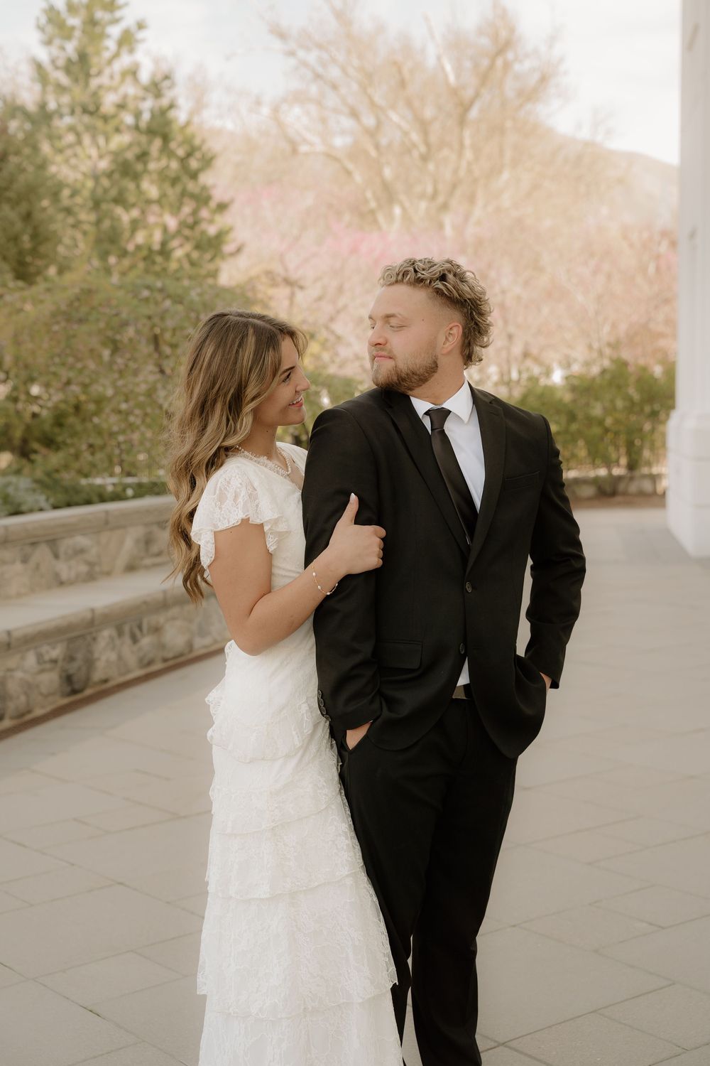 Bride in white ruffled dress and groom in black suit share a romantic moment outdoors with blooming trees.