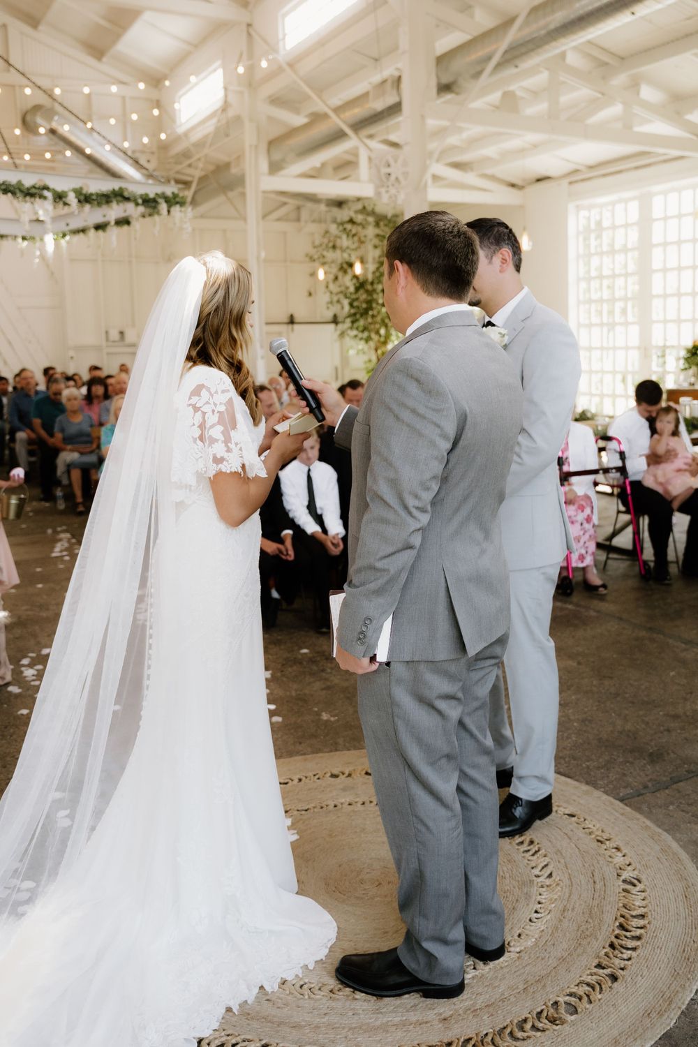 Wedding ceremony in progress inside bright industrial venue with string lights.