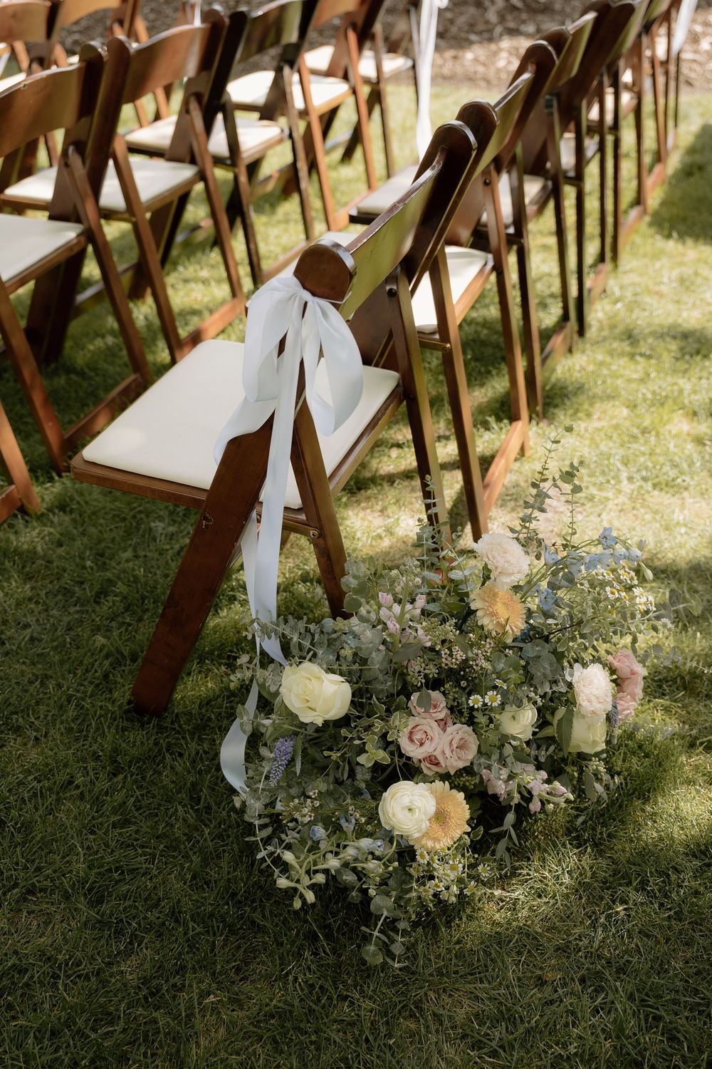 Floral wreath with pastel roses and greenery beside wooden folding chairs with white ribbon bows at an outdoor wedding ceremony.