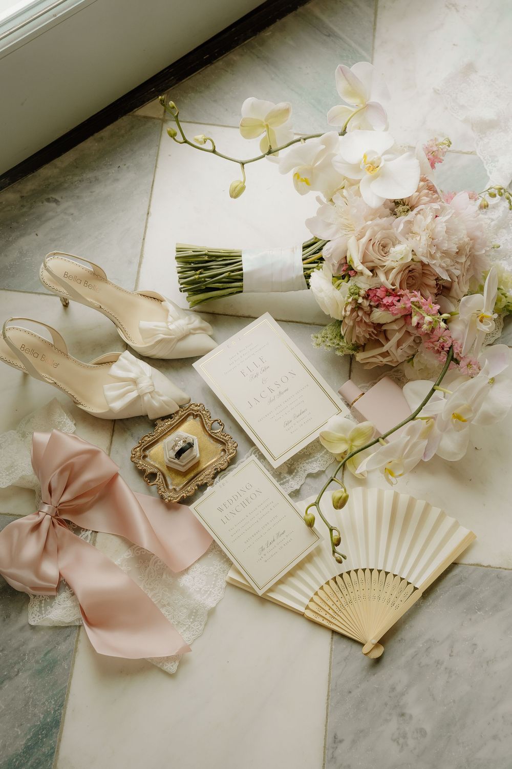 Elegant wedding flat lay with bridal heels, pink bow, floral bouquet, fan, invitation suite, and orchids on marble surface.