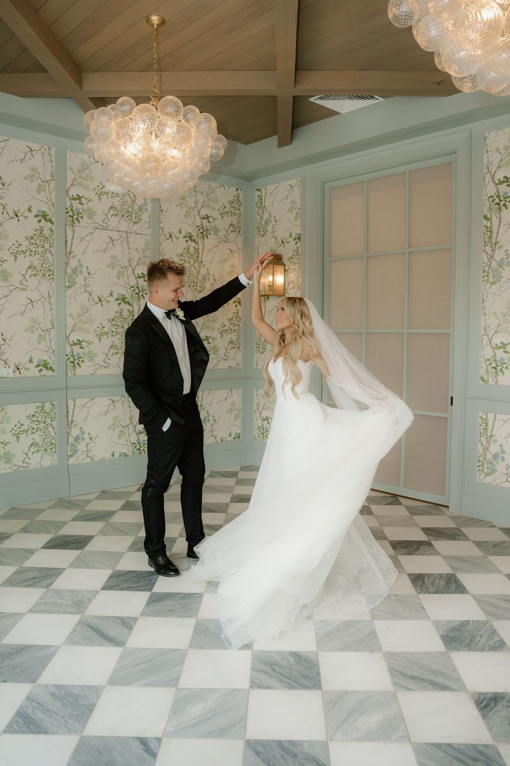 Bride and groom dancing in elegant room with checkered floors, floral wallpaper, and glass chandeliers.