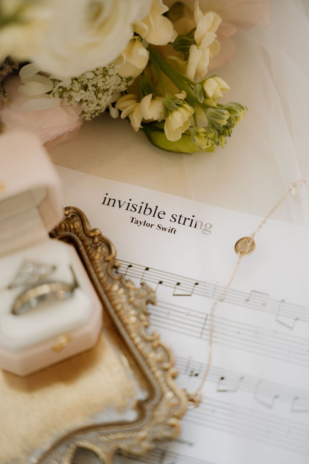 Wedding flat lay with rings, gold tray, necklace, Taylor Swift Invisible String sheet music, and white floral bouquet.