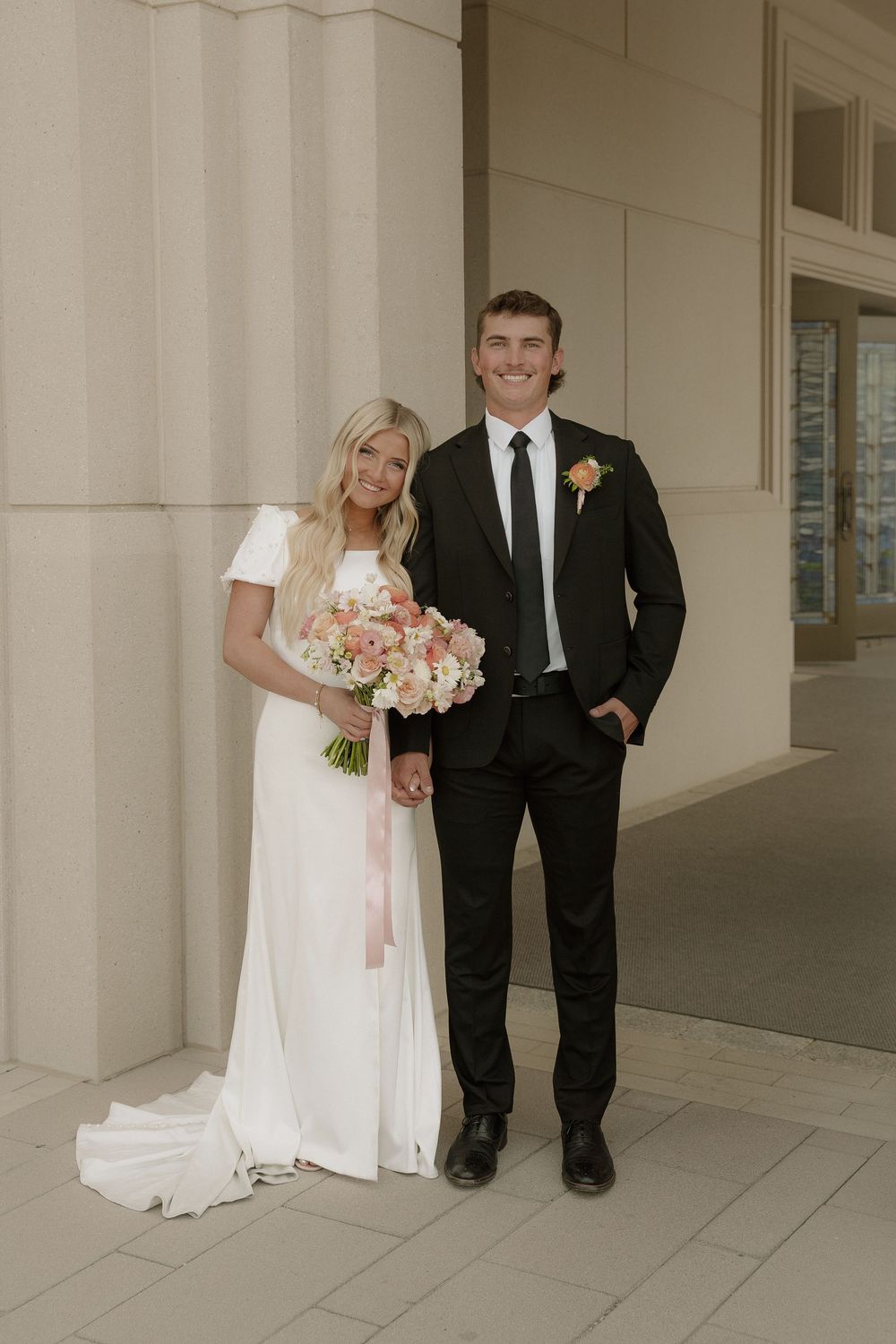 Bride in white gown holding pink bouquet stands with groom in black suit beside stone columns.